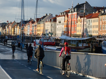 Copenhagen cycling This urban photograph captures cyclists riding along a dedicated bike path in Copenhagen during the afternoon in autumn. The main subject of the image is Copenhagen cycling, set against a backdrop of the city’s iconic architecture. Distinctive multicoloured historic buildings line the waterfront in the Nyhavn district, a well-known landmark in Copenhagen. Boats are moored along the canal, and the sunlight casts a warm glow over the city, emphasizing the vibrant architectural style typical of Copenhagen. The image effectively showcases cycling as an integral part of urban life in this city, while highlighting the architectural heritage and waterfront scenery unique to Copenhagen.